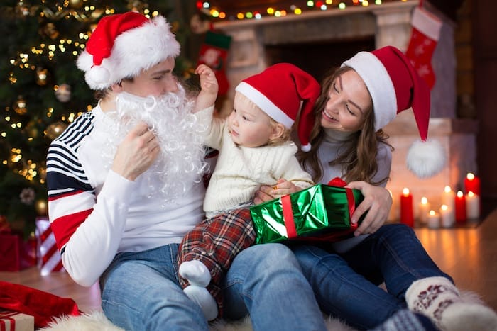 Surprised child boy looks at dad weared Santa Claus with fake beard sitting opposite Christmas tree