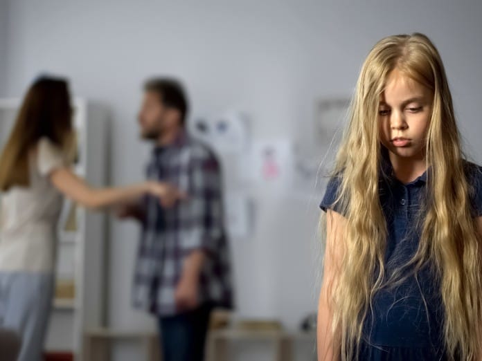 A young girl standing with her shoulders slumped and looking sad as her parents argue in the background.