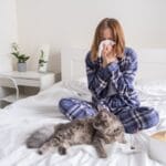 A homeowner laying in bed with her cat suffering from seasonal allergies symptoms such as a runny nose.