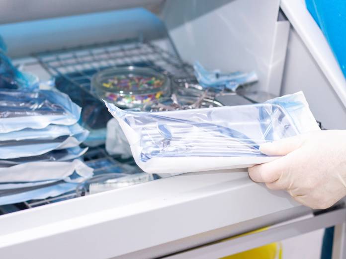 A shelf of medical devices in their secure packaging. A gloved hand picks up one of the packages to use.