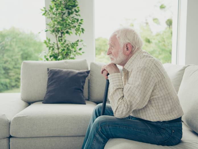 An old man is sitting by himself on the couch. He's resting his chin on his hands which tightly grip onto a cane.