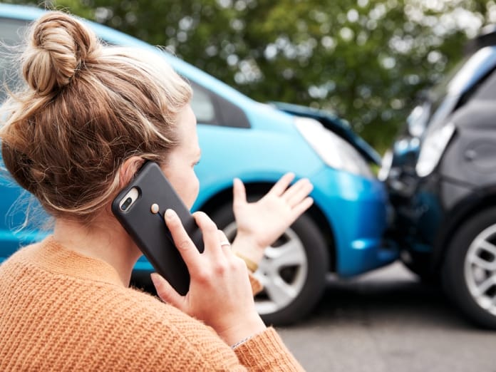 A woman is talking on a phone while looking at a car accident. She is wearing an orange sweater, and her hair is in a bun.