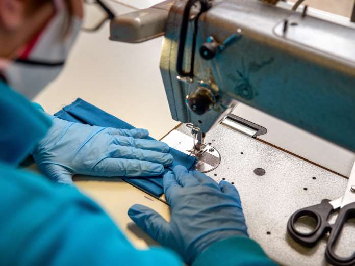 A close-up of a worker using an industrial sewing machine while wearing gloves and a face mask. There are scissors nearby.