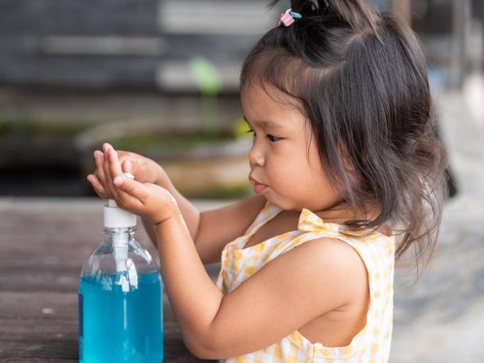 A little girl is seen pumping hand sanitizer onto her hands from a bottle filled with a light blue gel.