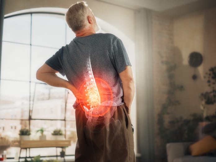 An older man wearing a gray shirt stands in his living room, holding his back. A digital overlay shows his spine in pain.