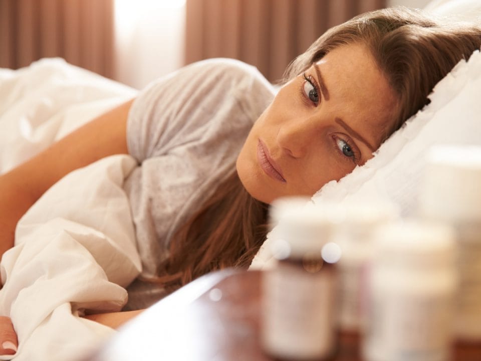 An unhappy young woman laying in bed and staring at a blurry medication bottle on her bedside table.