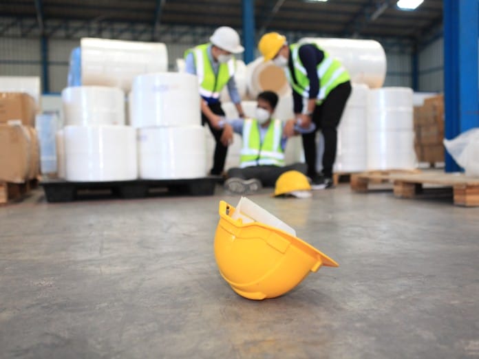 Focus on a yellow hard hat resting on a warehouse floor while two workers help a sitting worker in the background.