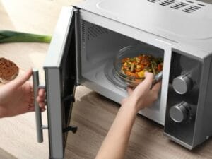 A person is placing a bowl of vegetables into a microwave. There are slices of bread on the counter next to the microwave.