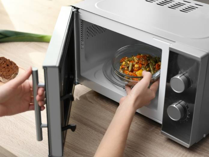 A person is placing a bowl of vegetables into a microwave. There are slices of bread on the counter next to the microwave.