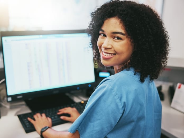 A young woman with dark, curly hair sits in a chair at a desk, typing on a keyboard. She smiles at the camera.