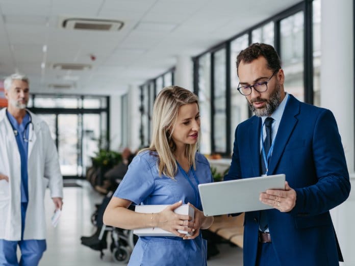 Two medical professionals, one in scrubs and the other in a suit, talking and looking at a tablet together.