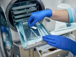 A medical lab professional standing next to a device autoclave. There is a new instrument on a tray in its packaging.