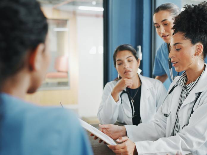 A doctor holds a clipboard in front of her as she talks to a group of medical professionals standing around her.