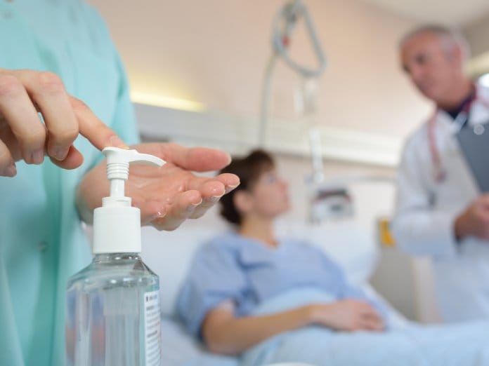 A healthcare worker squirts a shot of hand sanitizer into her hands. A doctor and patient speak behind her.
