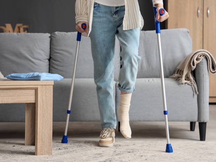 A person with a foot injury using crutches to stand. They are in front of their living room couch at home.
