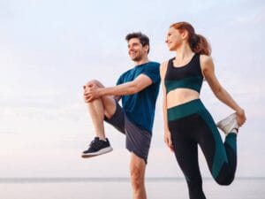 A man and a woman in workout clothing, smiling as they stretch their quads and hip flexors in front of a slightly pink sky.