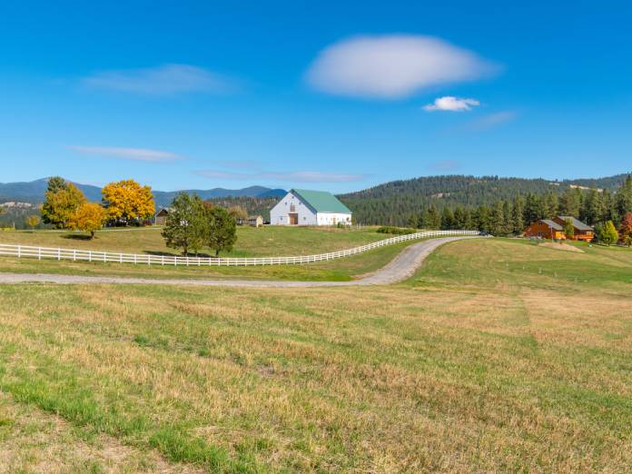 A large, white barn surrounded by a white picket fence sits in the middle of a large plot of hilly land.