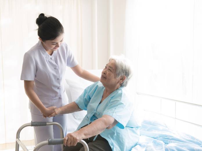 An older adult sits on the edge of their bed with one hand on a walker. A caregiver holds the individual's other hand.