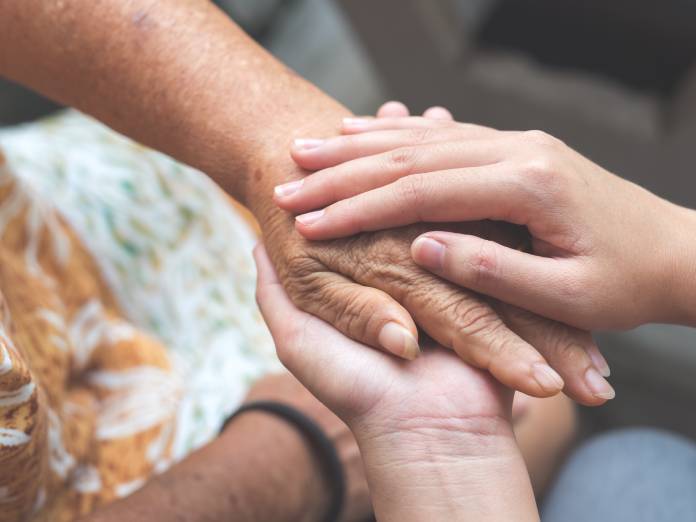 A young person's hands gently supporting and covering the hand of an elderly woman sitting next to them.