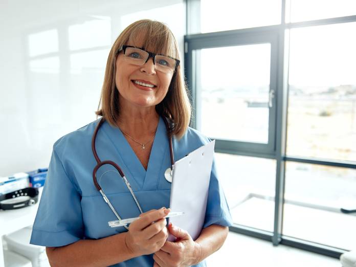 A female medical professional wearing blue scrubs has a stethoscope around her neck and holds a pen and clipboard.