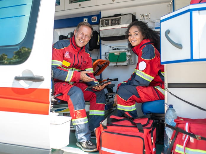 Two smiling paramedics wearing protective pants and jackets sitting in an ambulance with the doors open.