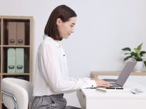 Woman sitting in a white leader chair with an upright straight position working on a laptop placed over a desk.