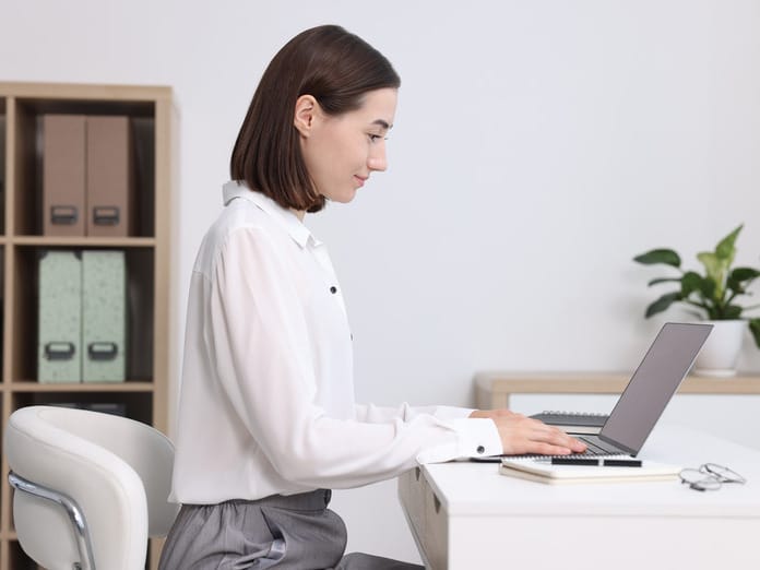 Woman sitting in a white leader chair with an upright straight position working on a laptop placed over a desk.