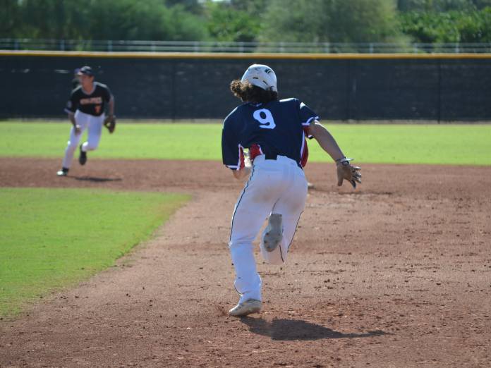 The back of a baseball player sprinting to a base on a field, one of his teammates in the background.