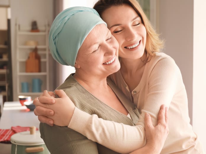 Two women, one of whom is wearing a scarf on her head, hugging. Behind them is a kitchen counter with a pot and bowls.
