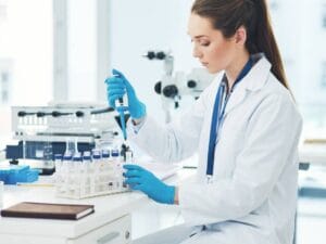 A female pharmaceutical lab technician working closely with a sample. She's wearing blue latex gloves to handle the sample.