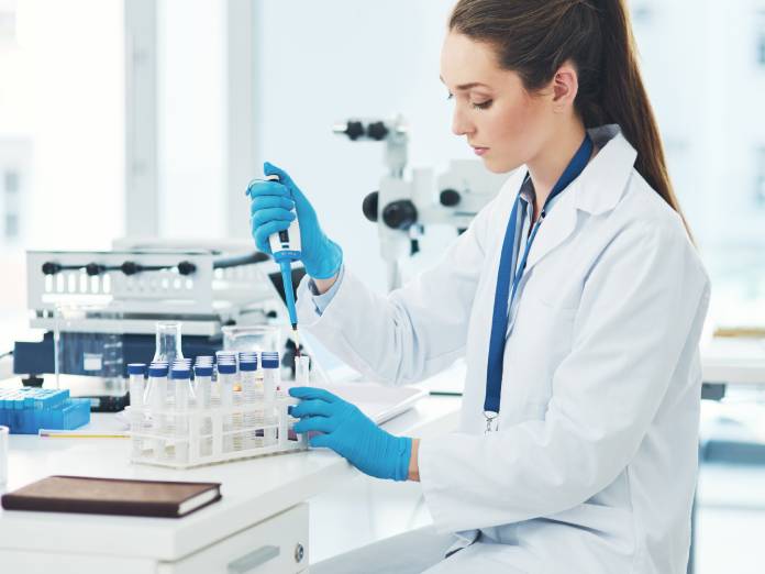A female pharmaceutical lab technician working closely with a sample. She's wearing blue latex gloves to handle the sample.