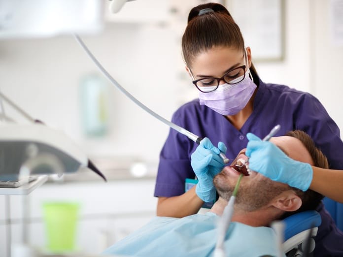 A female dentist in purple scrubs and a purple mask using stainless steel tools to clean a male patient's mouth.