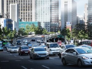 A close-up of a traffic jam in the center of a large modern city. There are tall buildings and trees in the background.