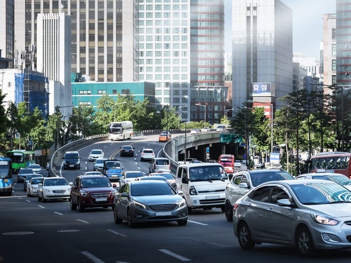A close-up of a traffic jam in the center of a large modern city. There are tall buildings and trees in the background.