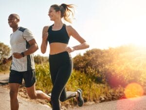 A man and a woman in black and gray workout gear smiling and laughing while running outside on a sunny day.