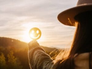 A woman standing outside and holding up a smiley face cutout in front of the sun as it sets behind a hill.