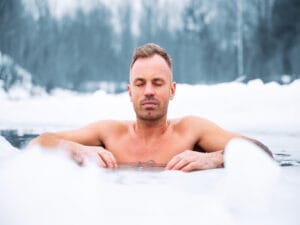 A close-up view shows a man in the cold water surrounded by ice. He is closing his eyes and concentrating.