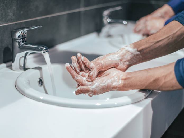 A close-up of a person with rolled up sleeves washing their hands at a public restroom sink next to another person.