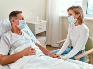 A younger person visiting a slightly older person in a hospital bed. They are both wearing blue face masks.
