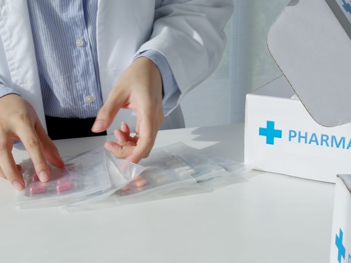 A pharmacist in a white coat is packing up orange pills in a bag. A box on the counter says "PHARMACY" in blue letters.