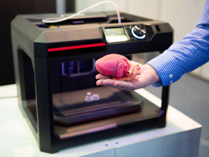A medical professional holding a 3D printed heart in their right hand. The 3D printer is on a table behind them.