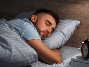 A young man sleeps on striped gray sheets in a bed with a wooden headboard. A clock sits on the nightstand.