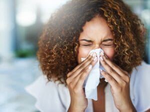 A woman with short, tightly curled hair scrunches her eyes as she sneezes into a white tissue.