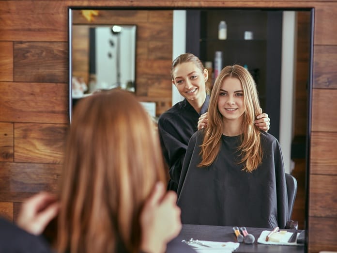 A hairstylist and a client look at one another in a mirror. They smile while the stylist touches the client's hair.