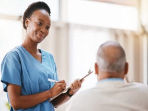 A woman wearing blue scrubs and a stethoscope looks down at a seated older man while holding a pen and a clipboard.