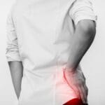 A close-up, rear view of a man holding his hip. A red glow illuminates his hip. He wears a white shirt and dark pants.