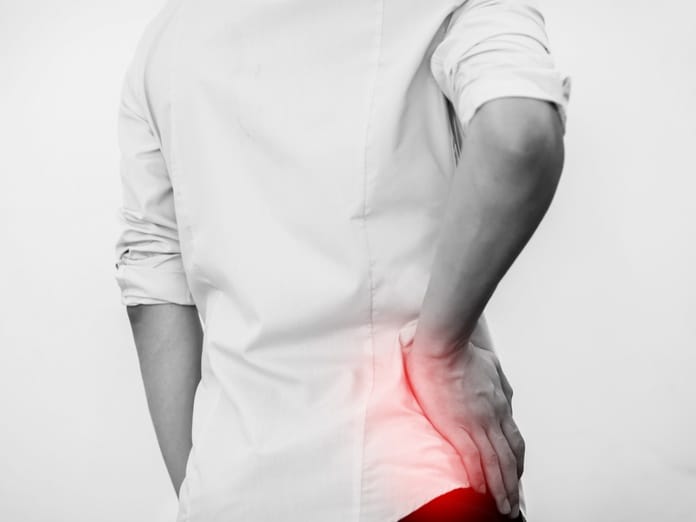A close-up, rear view of a man holding his hip. A red glow illuminates his hip. He wears a white shirt and dark pants.