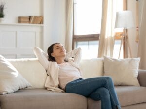 A woman sits on her sofa at home with her eyes closed. She relaxes with her legs crossed and her hands behind her head.