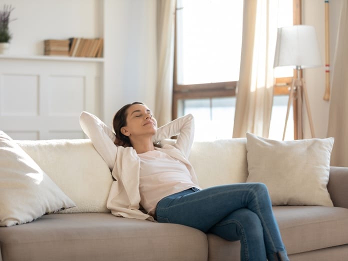 A woman sits on her sofa at home with her eyes closed. She relaxes with her legs crossed and her hands behind her head.