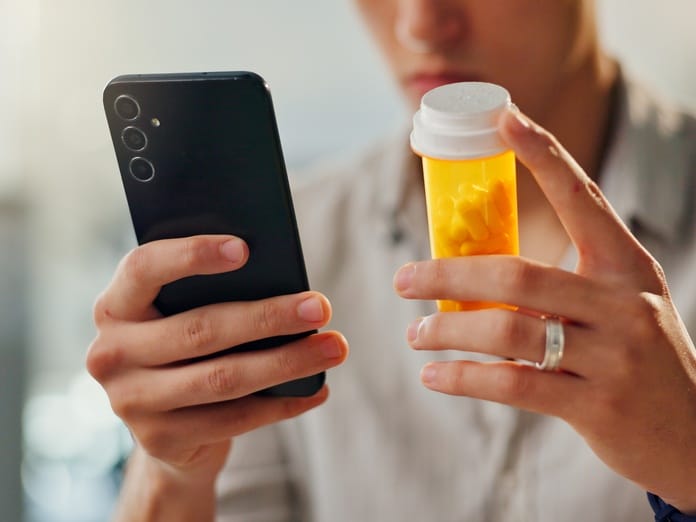 A man wearing a black watch holding a smartphone in one hand and an orange medication bottle in the other hand.